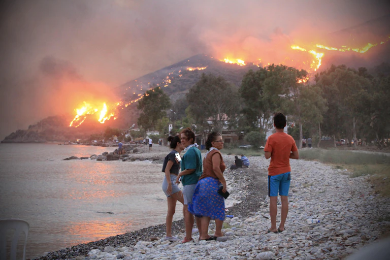 Four people stand on a stony beach watching fires burn on a hillside in the background