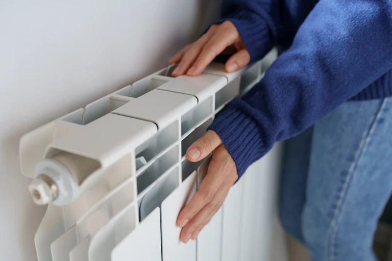 woman hands, wearing blue jumper, touching radiator against white wall