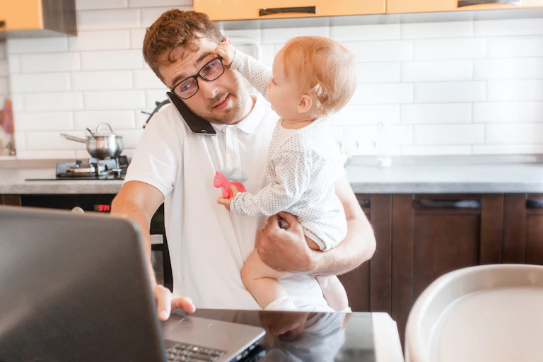 young man at a laptop holding a baby who is hitting him in the face