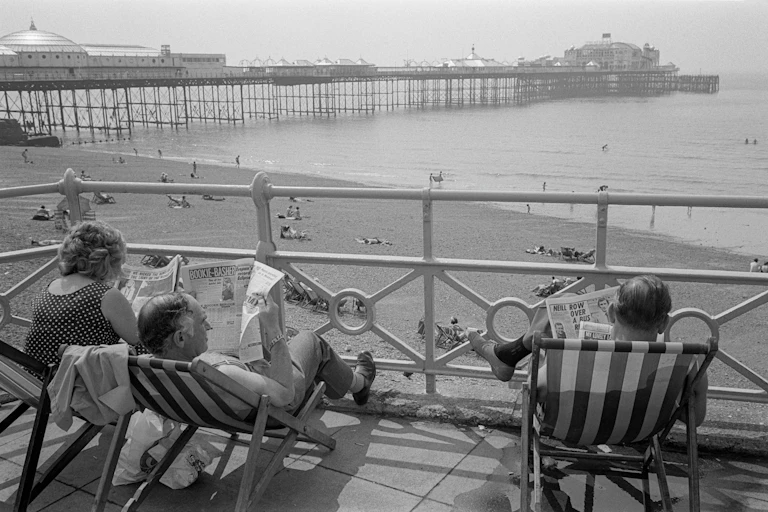three people sat in deckchairs on Brighton seafront reading newspapers, black and white