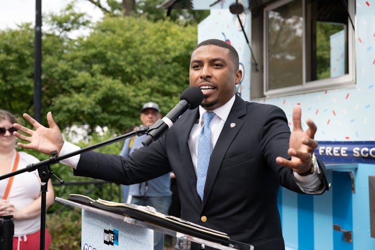 A man dressed in dark suit and light blue tie gestures while speaking outdoors at a podium