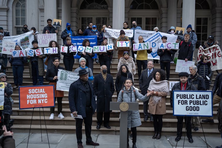 Activists stand on steps holding colorful signs while a woman stands in front of them speaking from a lectern.