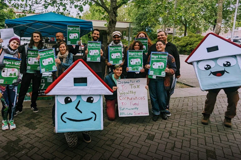 A group of people hold green signs next to two people dressed up as anthropomorphic houses.