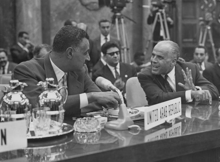Two men in suits sit behind a desk chatting.