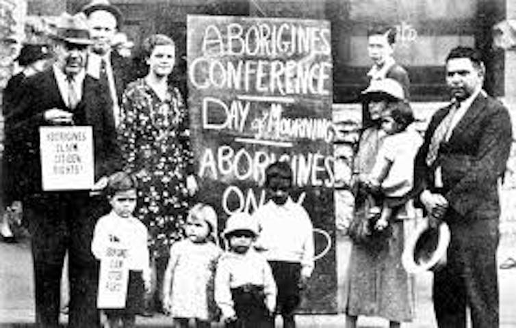 Adults and children gathered around a Aboriginal Day of Mourning banner.