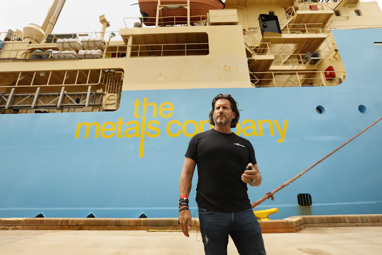 A man holding a nodule from the deep sea stands on the dock with a ship labelled The Metals Company behind him.