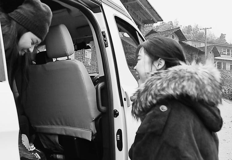a girl in a beanie is getting out of a van, with her twin looking at her, smiling, in a parka