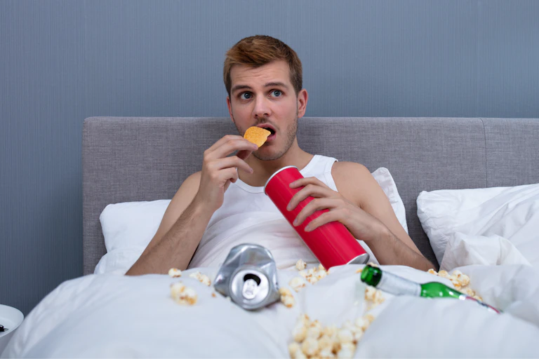 A man is eating snacks in a dirty bed.