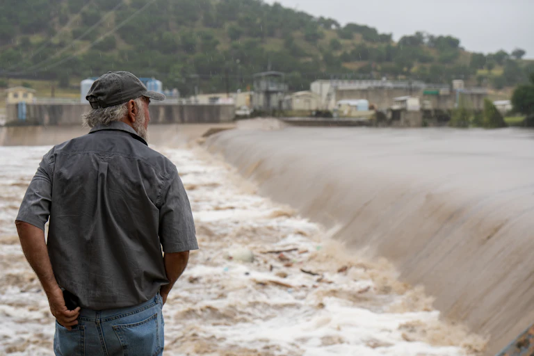 A man in a baseball cap watches flood water pouring down a river.