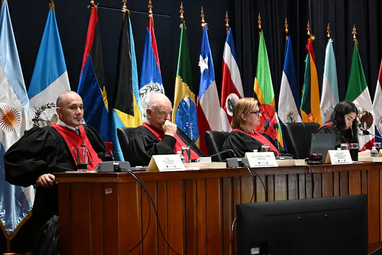 four judges sit at a table with flags behind them.