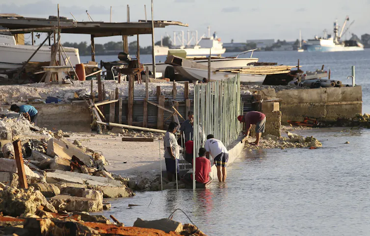 people stand in water and work on a wall with boats around them.