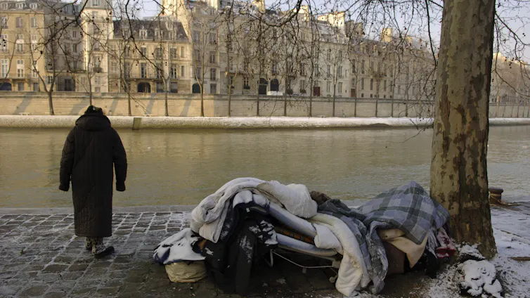 A woman stands by a river next to a homeless person's bedding.