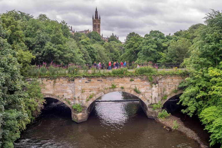 stone bridge over river, green trees either side, people walking