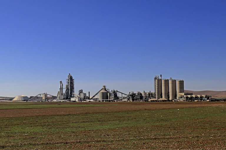 A large cement plant on flat land under a blue sky is seen in the distance.