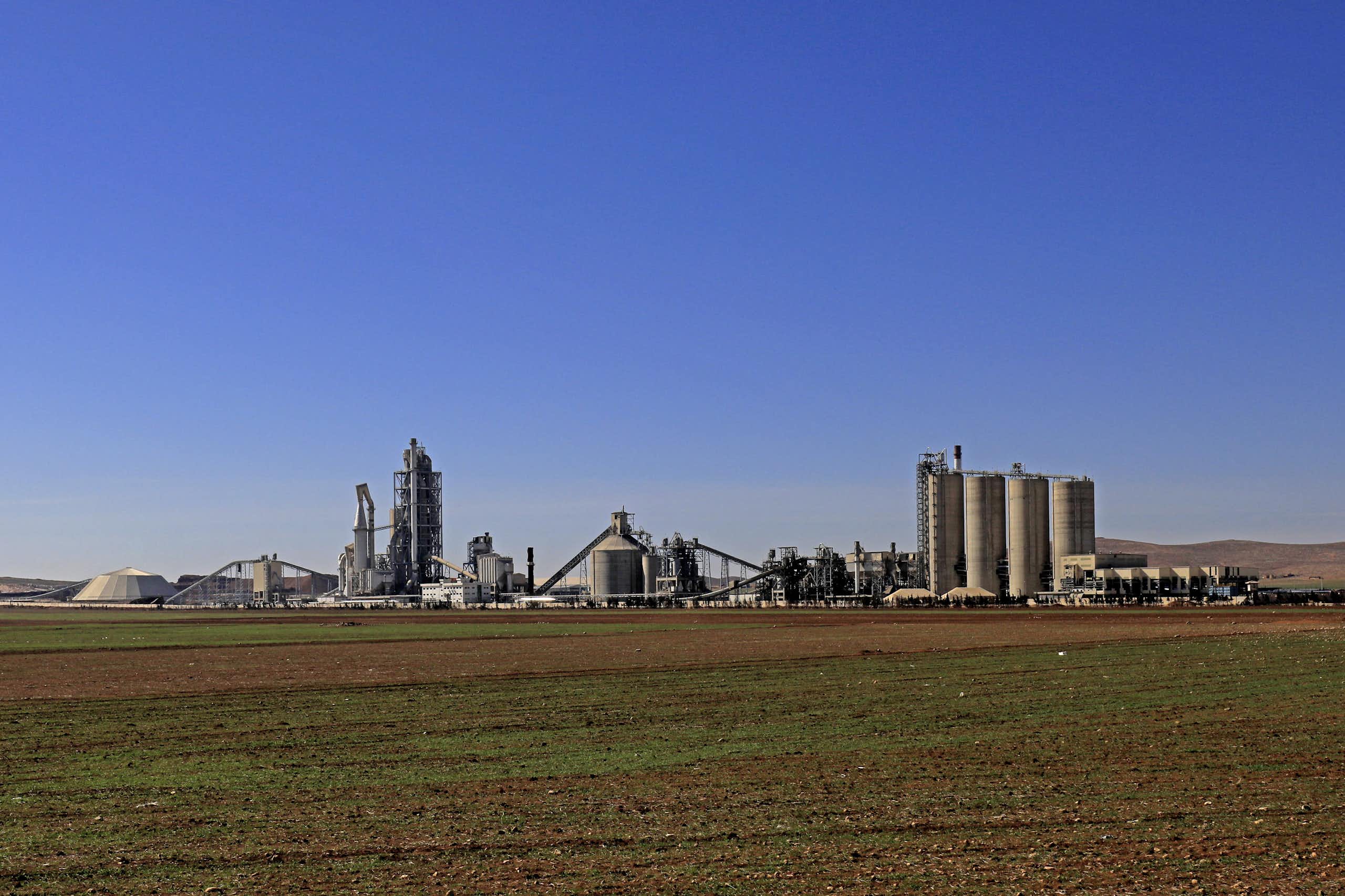 A large cement plant on flat land under a blue sky is seen in the distance.