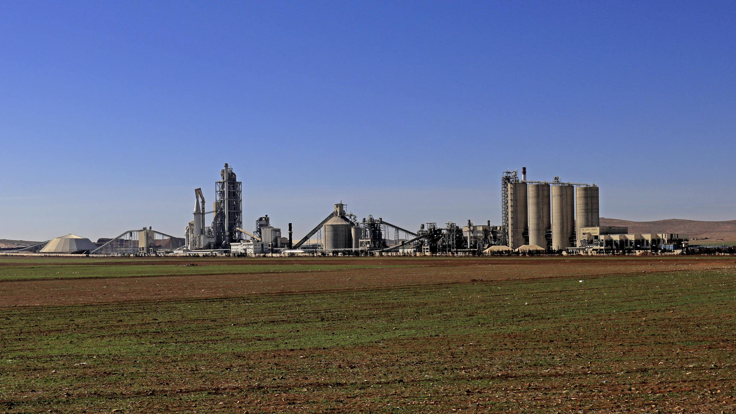 A large cement plant on flat land under a blue sky is seen in the distance.