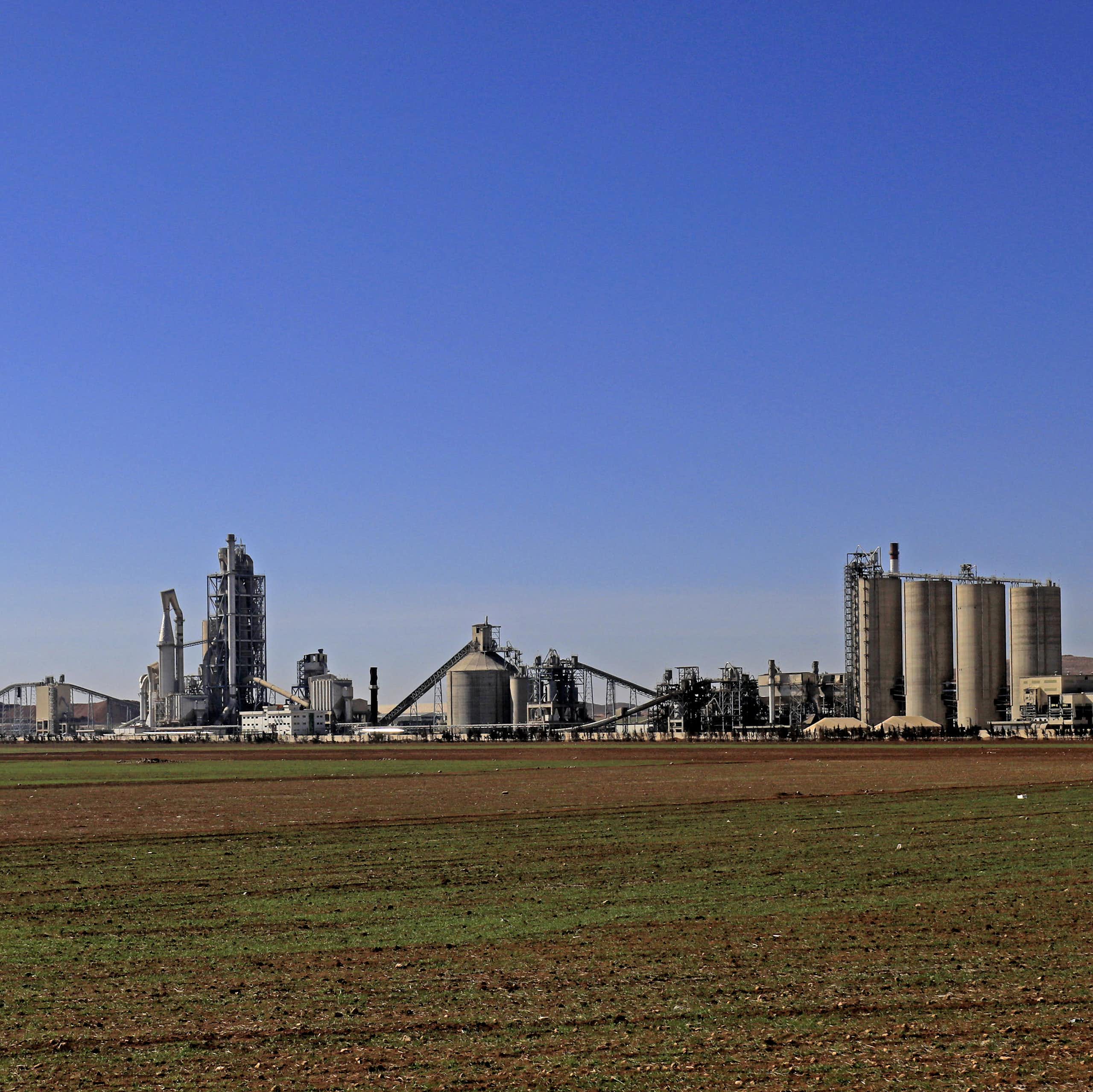 A large cement plant on flat land under a blue sky is seen in the distance.