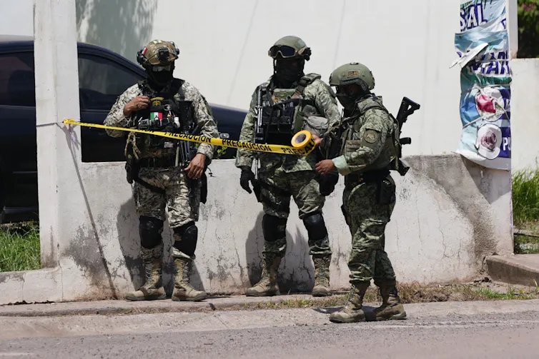 Three heavily armed soldiers carrying weapons stand in a street; one of them holds yellow police tape.