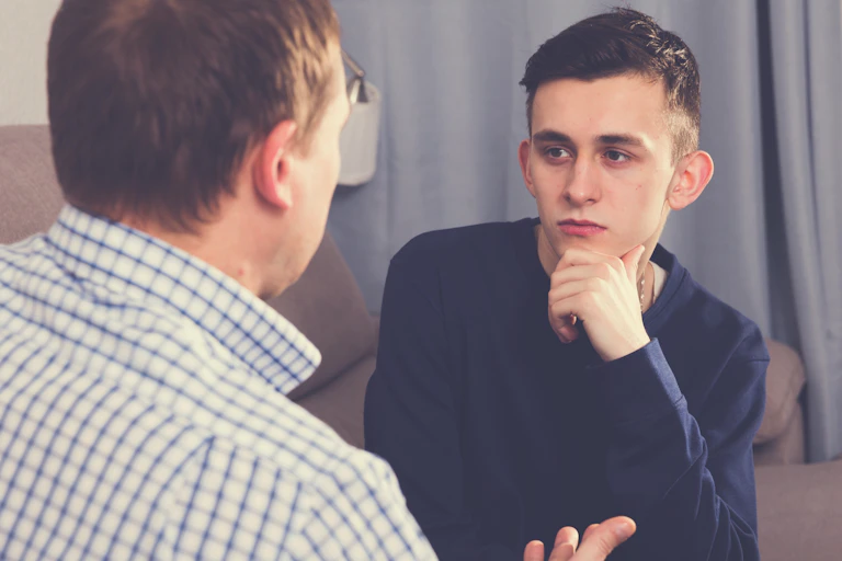 Un homme et son fils adolescent sont assis sur un canapé et ont une conversation sérieuse. La caméra est placée derrière le père, de sorte que seul le visage du fils est visible.
