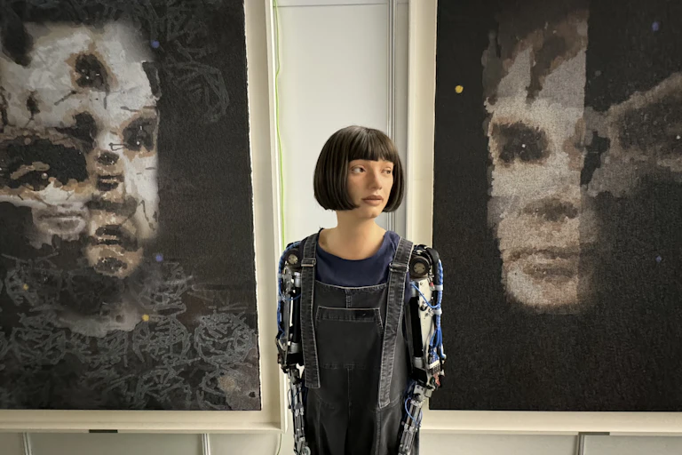 A robot looking like a woman with a bob haircut stands in front of two large black and white paintings