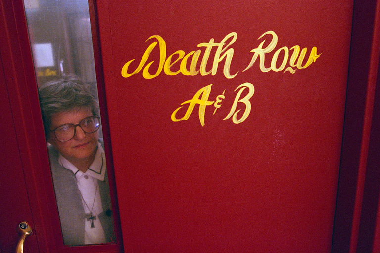 A woman with gray hair peers out from a narrow window next to a red door that says 'Death Row A&B.'