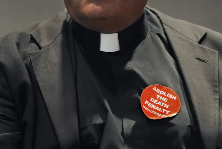 A close-up photo of the chest of a man wearing a priest's collar and a red sticker that says 'abolish the death penalty.'