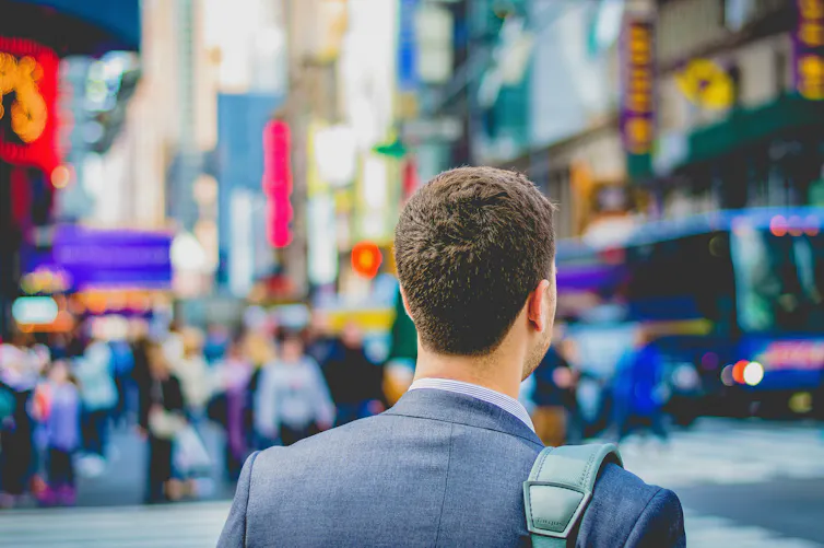 from the back, shoulders of a young person w short hair in a suit, a bag strap is visible, the foreground is a busy city