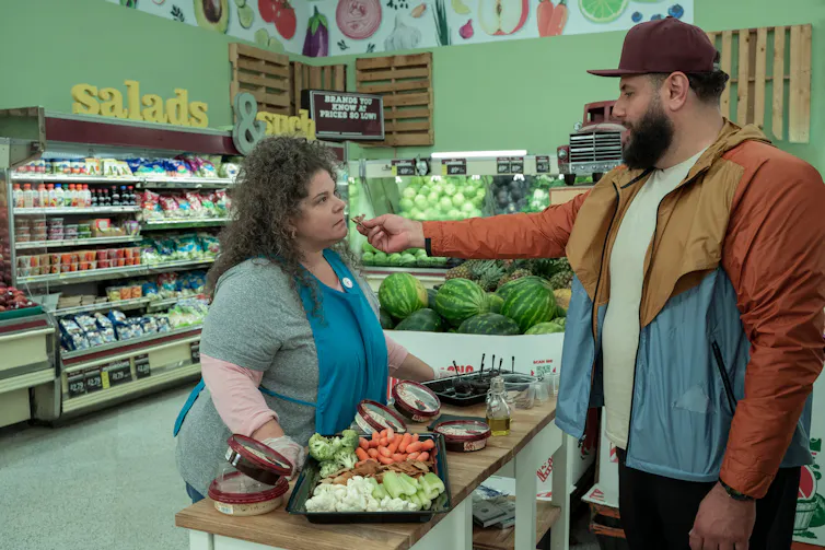 a man is feeding a woman in a grocery store. behind them is produce
