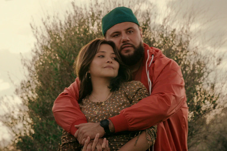 a man wraps his arms around a woman, both face forward. behind them is an olive tree.