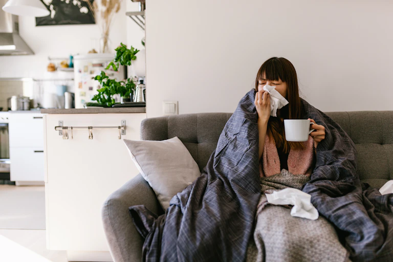 A woman sits on a couch in her home wrapped in a blanket and using tissues.