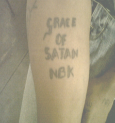 A human arm with a black tattoo on the inside with the words Grace of Satan NBK.