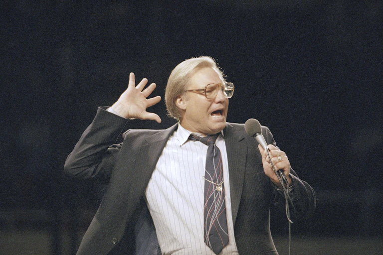 A man in a gray suit and tie gestures with his hand as he speaks into the microphone.