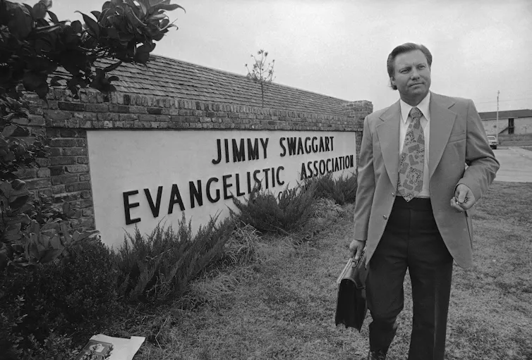 A man holding a briefcase in one hand stands next to a sign on a wall that says, 'Jimmy Swaggart Evangelistic Association.'