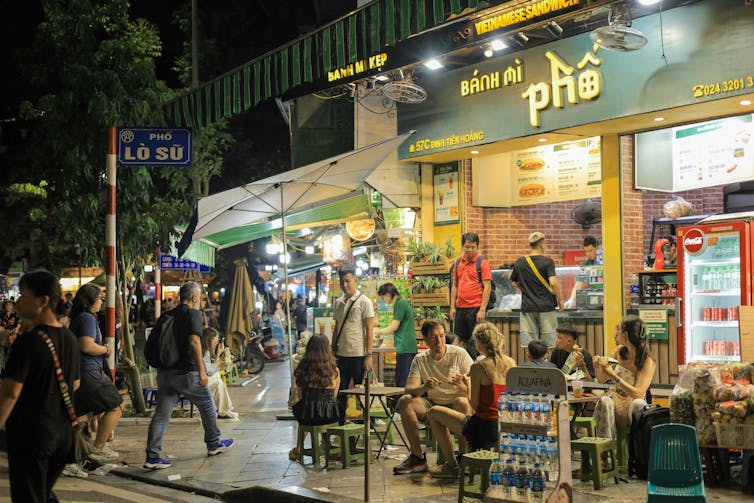 Escena callejera con personas sentadas comiendo en la terraza de un puesto de comida