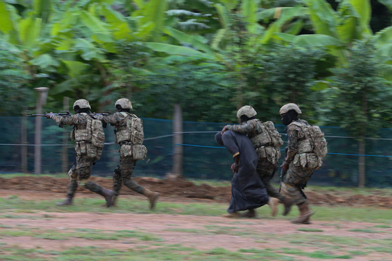 People in combat uniform, moving quickly. One has a hand on a person who is dressed in a robe and is bent over.