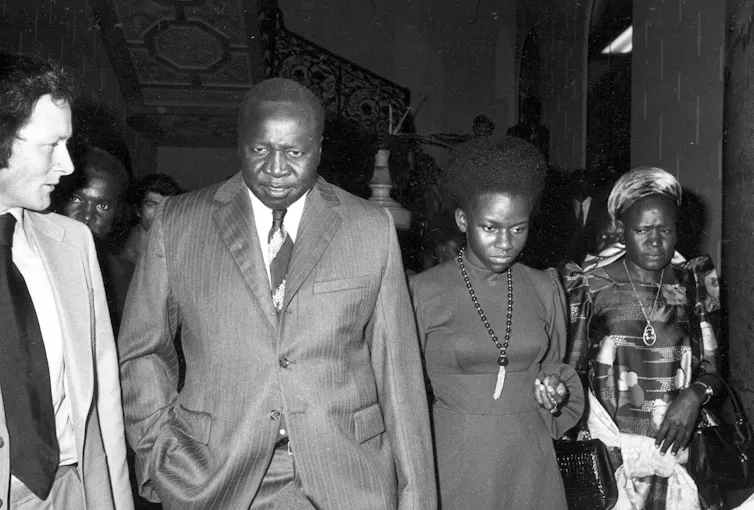 A Black man in a suit walking with a woman and another dressed in traditional African attire.