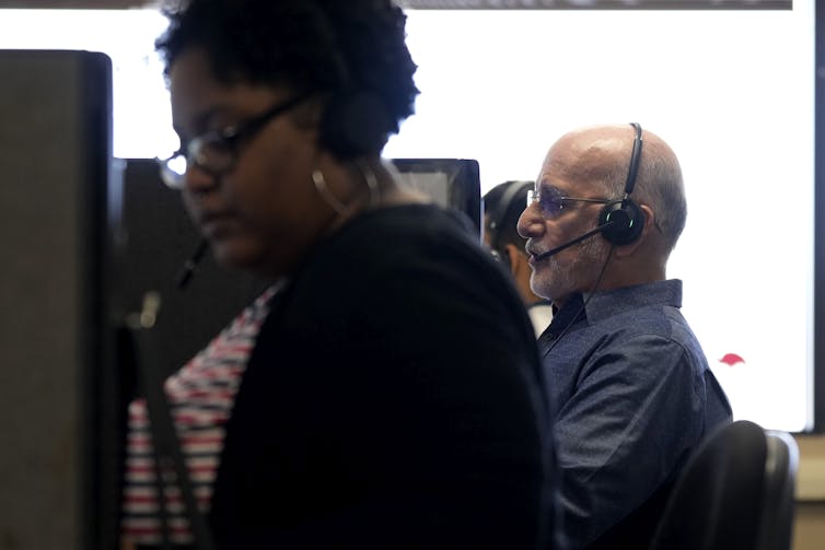 Two people sit at their own cubicles with headsets on