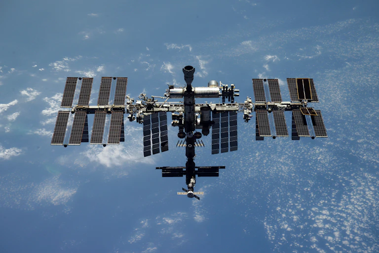 The ISS orbiting over Earth. It has two arrays of large solar panels and a cylindrical chamber in the center.