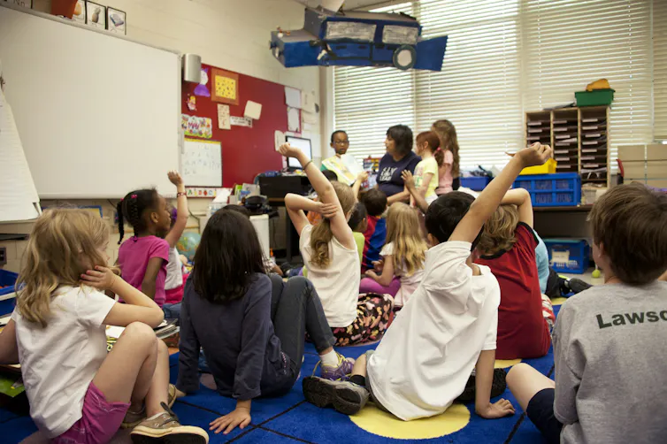 Children in a classroom close to a teacher reading a book.