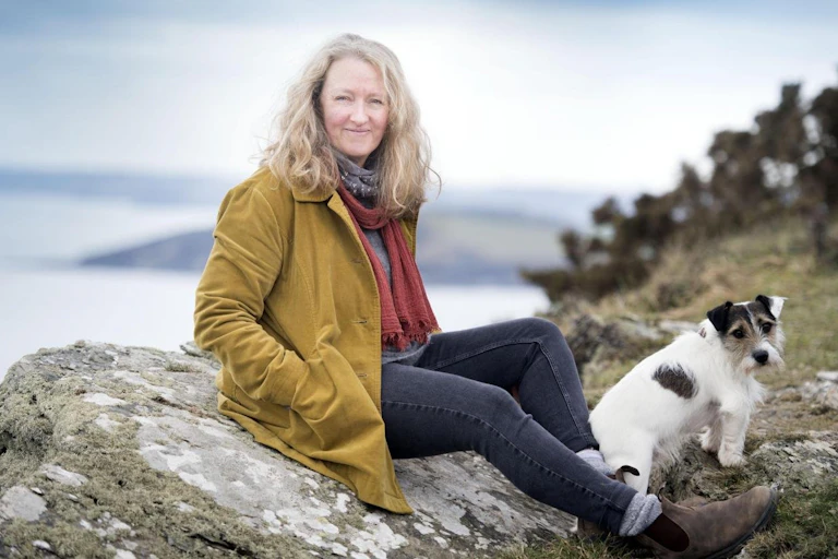 Woman in mustard-coloured jacket sits on a rock next to the sea, accompanied by a dog