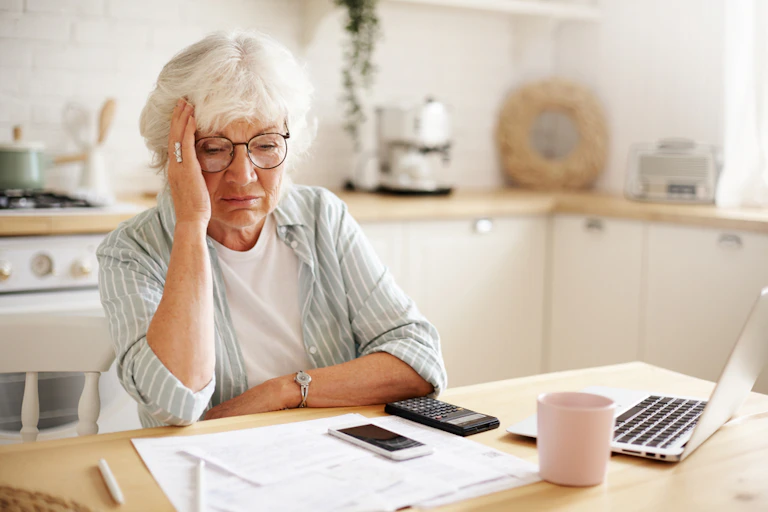 Elderly woman sitting at kitchen table with laptop, hand holding head