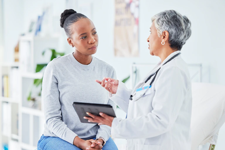 A woman is sitting on a table and listening to another woman wearing a white coat and holding a tablet.