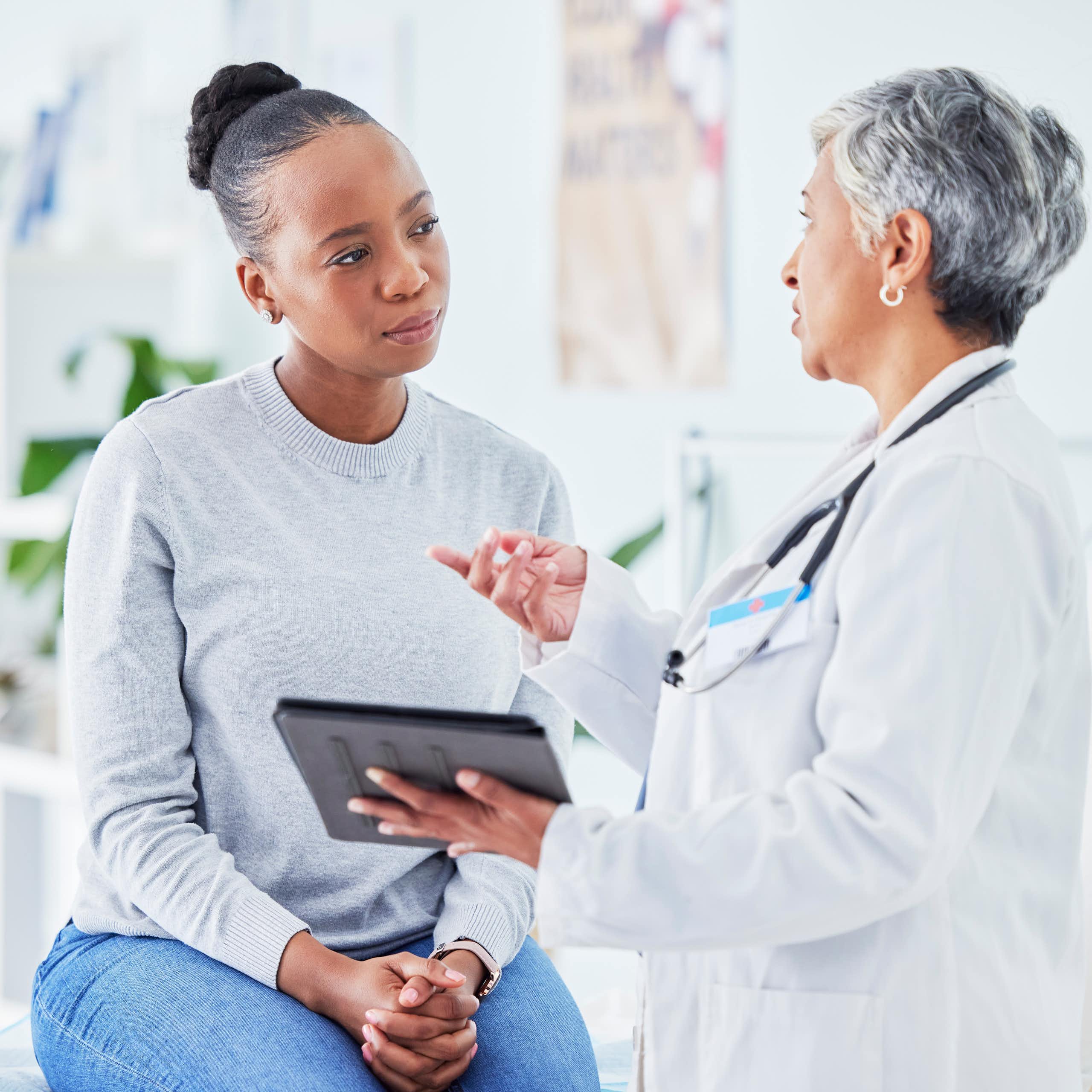 A woman is sitting on a table and listening to another woman wearing a white coat and holding a tablet.