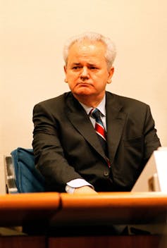 A middle-aged man sits in a courtroom dressed in a suit