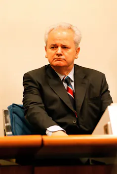 A middle-aged man sits in a courtroom dressed in a suit