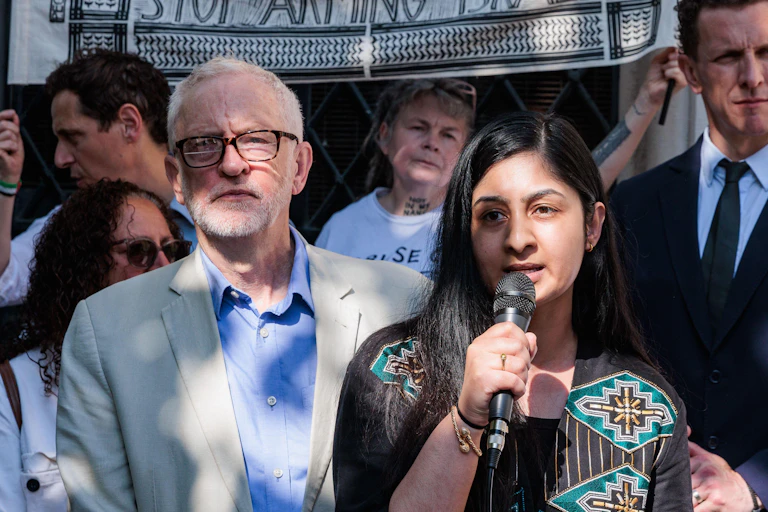 Corbyn stands next to Sultana, who is speaking into a microphone
