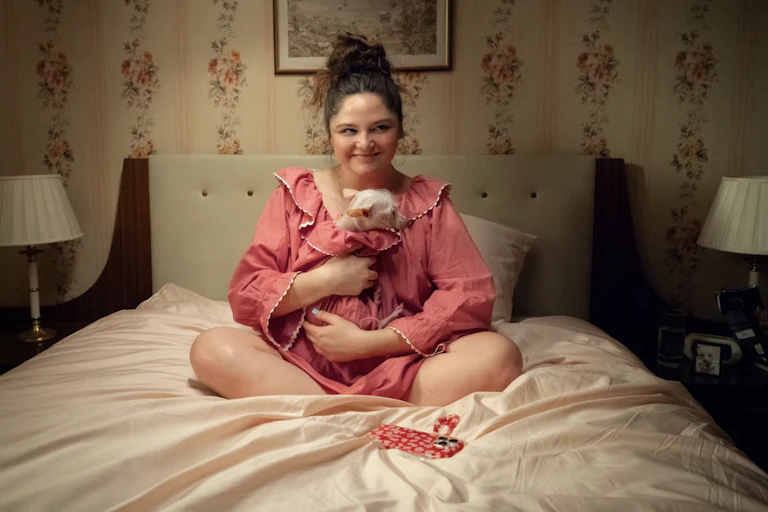 A young woman sitting on a bed smiling holding a small ratty-looking dog.