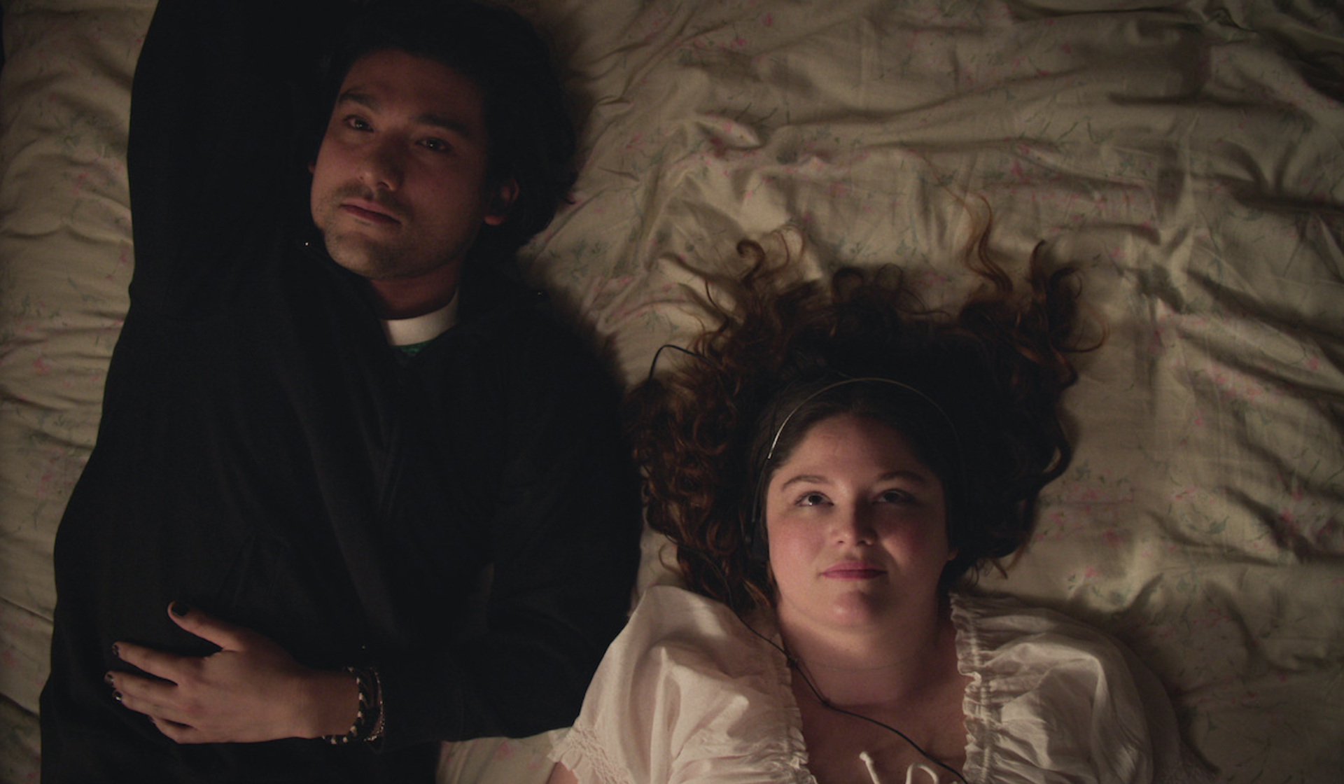 A young couple lying on a bed facing up to the ceiling, looking thoughtful.
