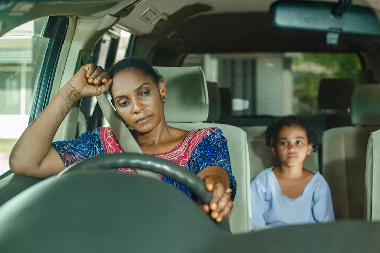 A woman behind the wheel of a car looking stressed as a child sits in the back seat.