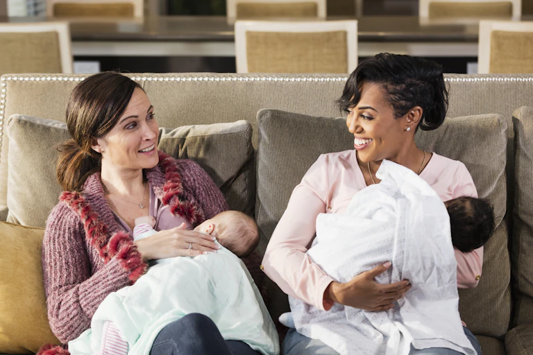 Two women holding babies sit next to each other on a couch and talk.